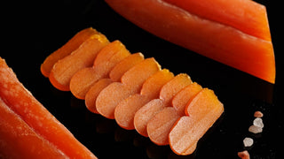 Orange bottarga, whole and in thin slices, on a black surface, with a few grains of pink salt.