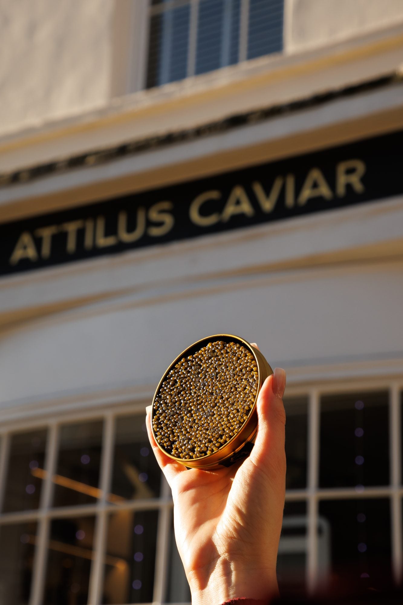 A hand holds up an open tin of caviar in front of a shopfront whose sign reads Attilus Caviar.