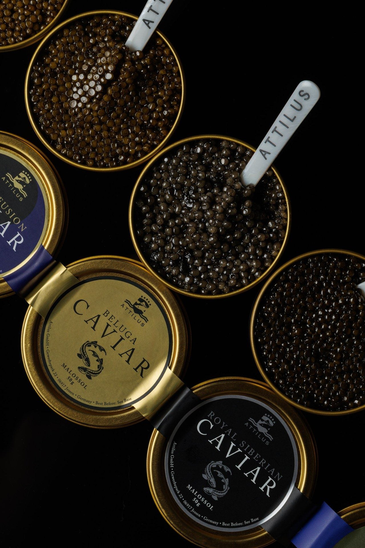 Open tins of caviar with Attilus mother-of-pearl spoons on a black surface, beside two closed tins whose lids read Beluga and Royal Siberian.