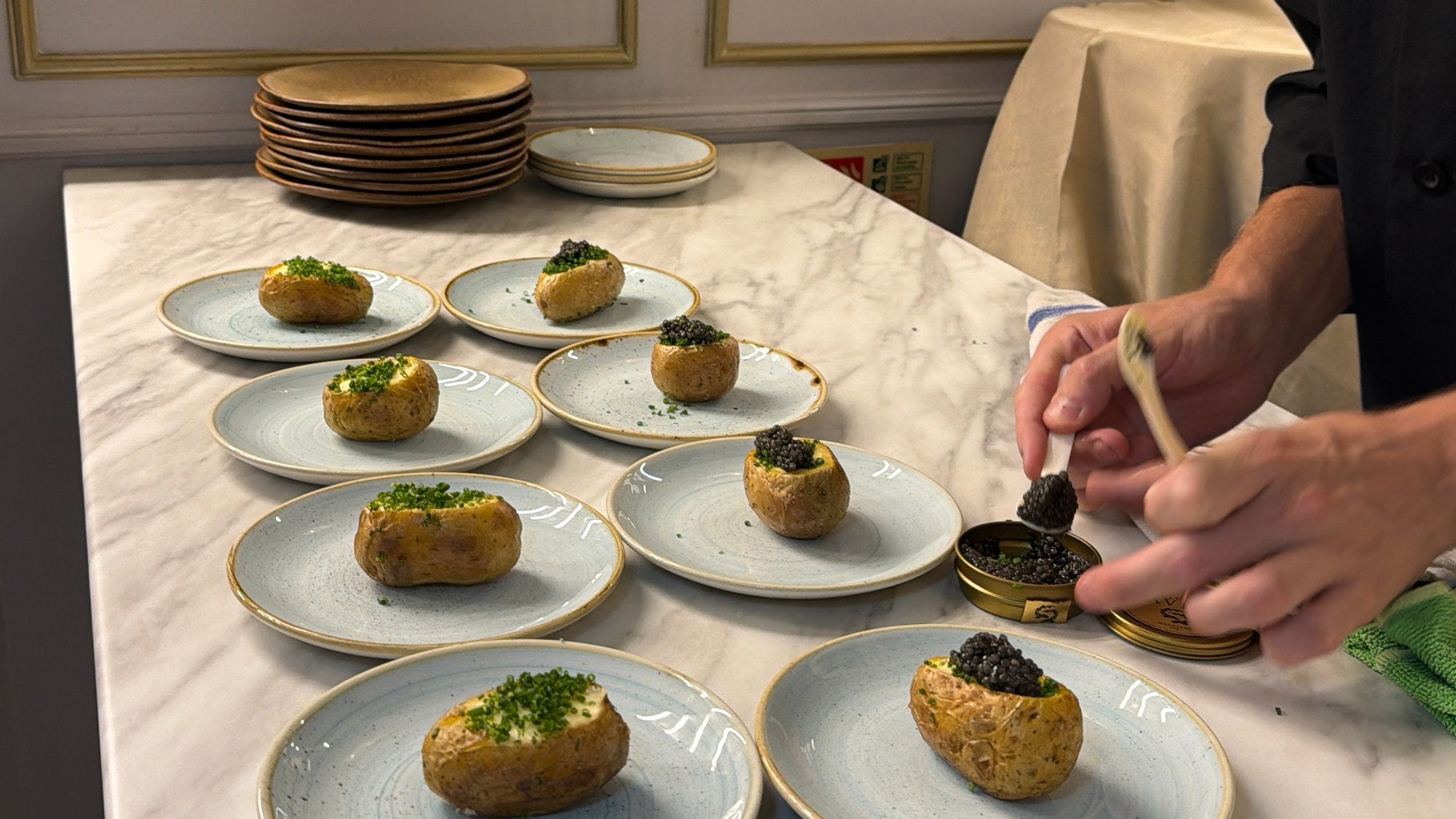 A chef's hands spooning caviar from an open Attilus tin onto potatoes topped with chives, on pale blue plates along a marble counter.