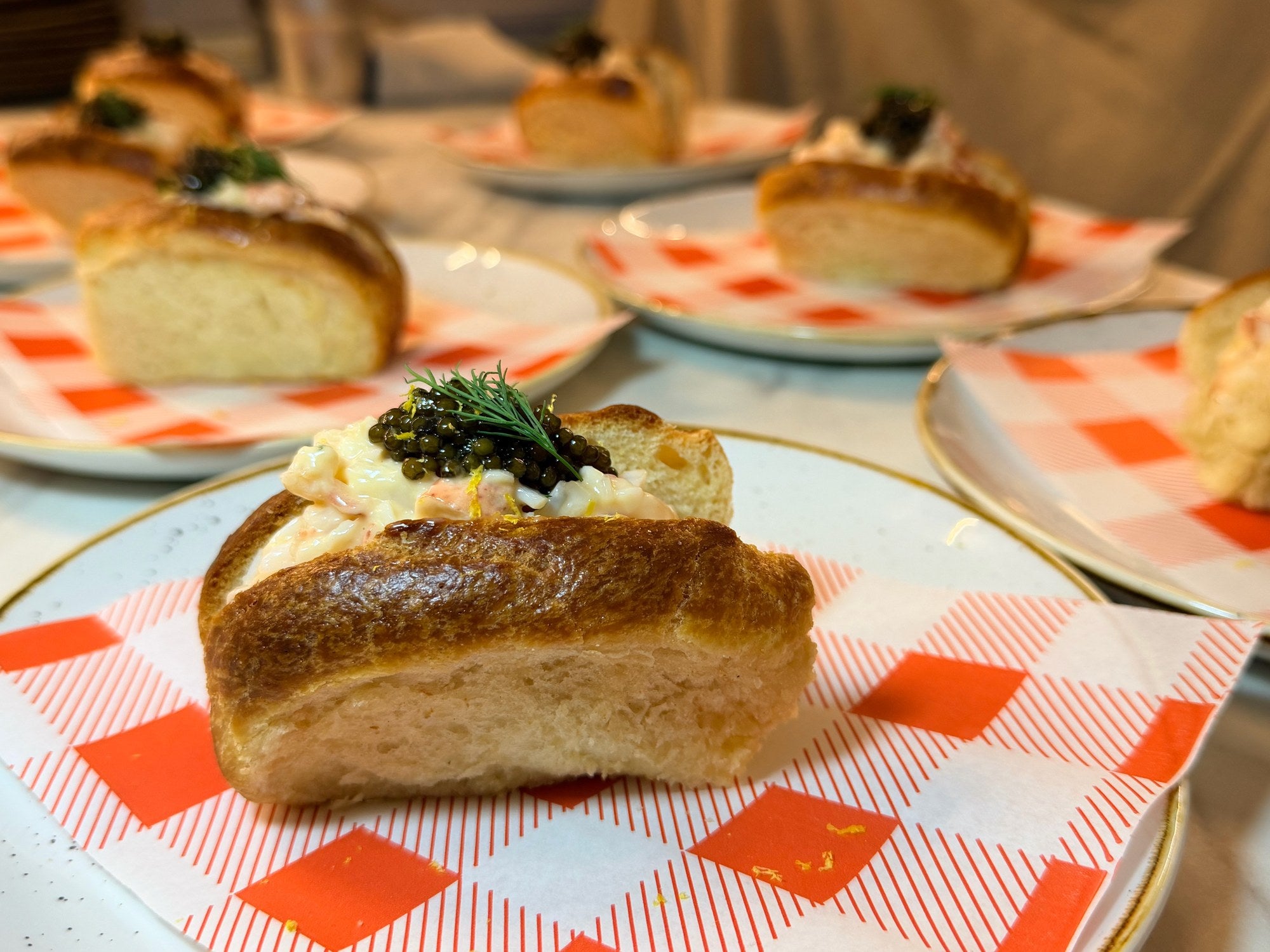 A lobster roll in a brioche bun on red and white gingham paper, topped with Amur Fusion caviar, a sprig of dill and lemon zest, with more rolls behind.