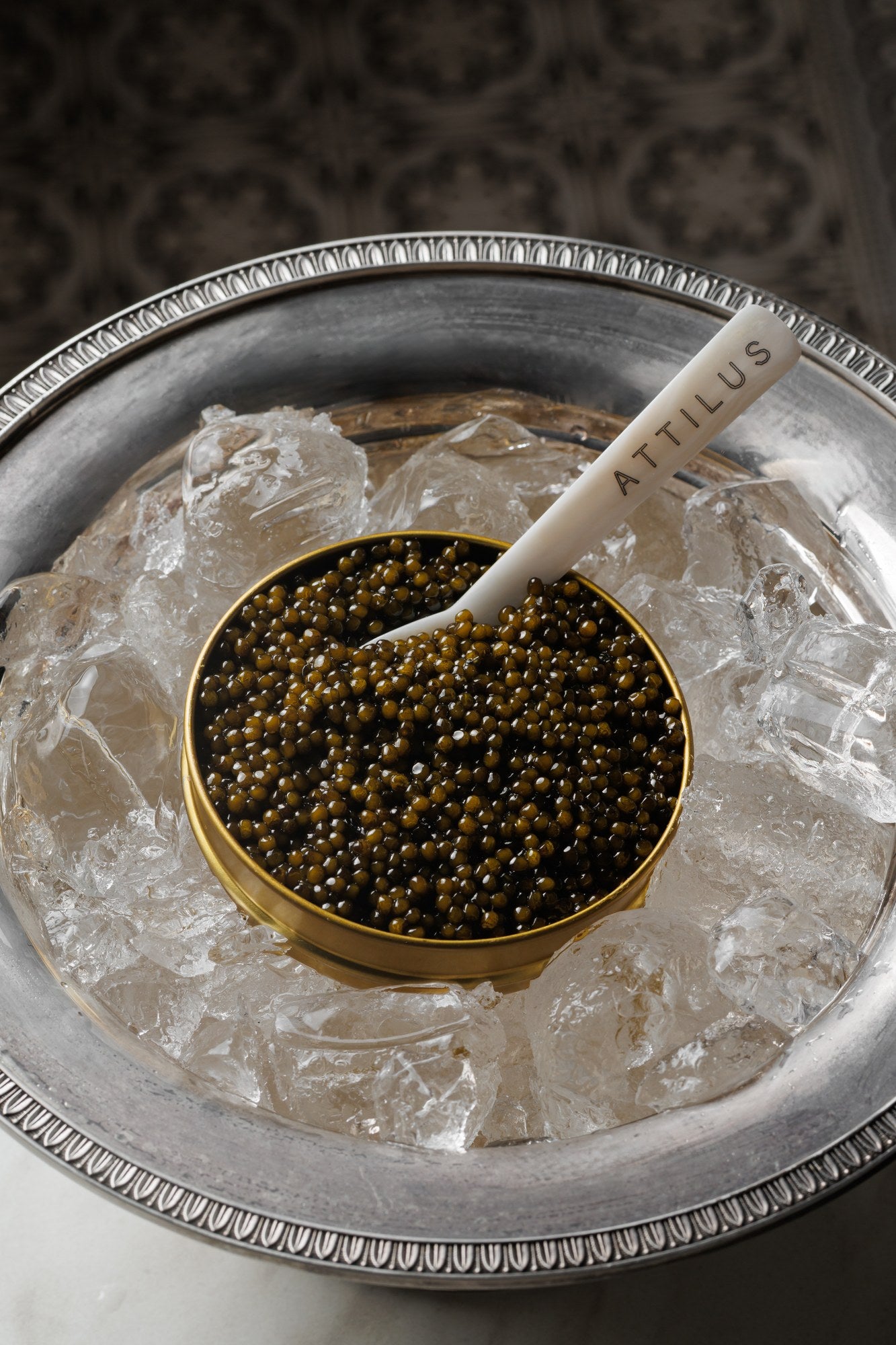 An open tin of caviar set among ice cubes in a silver dish, with an Attilus mother-of-pearl spoon.