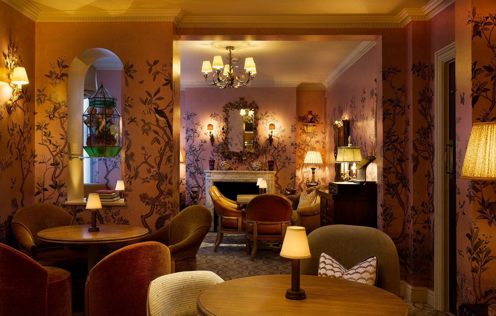 The Delany Drawing Room: hand-painted wallpaper of birds and branches, velvet armchairs around small tables, table lamps and a marble fireplace.
