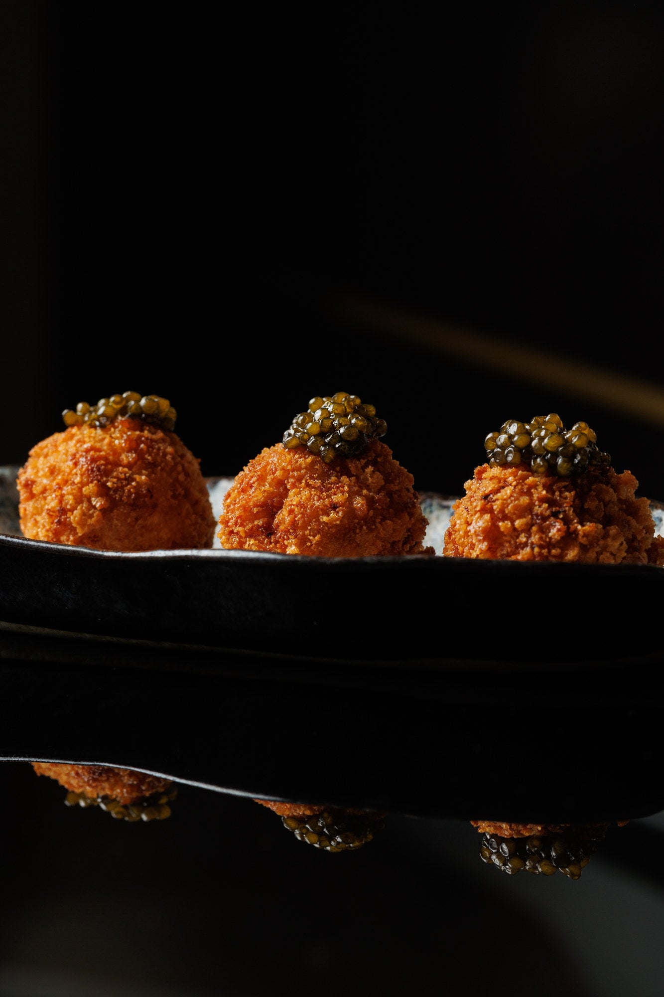 Three crisp golden croquettes in a row on a dark ceramic plate, each topped with a spoonful of dark caviar, against a black background.