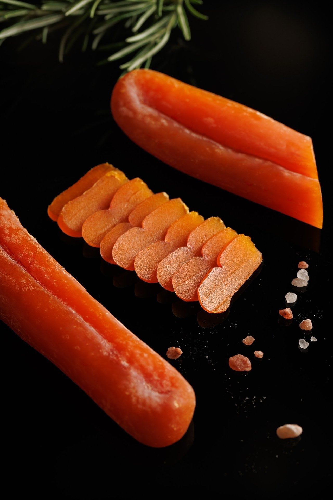 Two whole pieces of orange bottarga, one cut at the end, with a fan of thin slices between them on a black surface, grains of pink salt and a sprig of rosemary.
