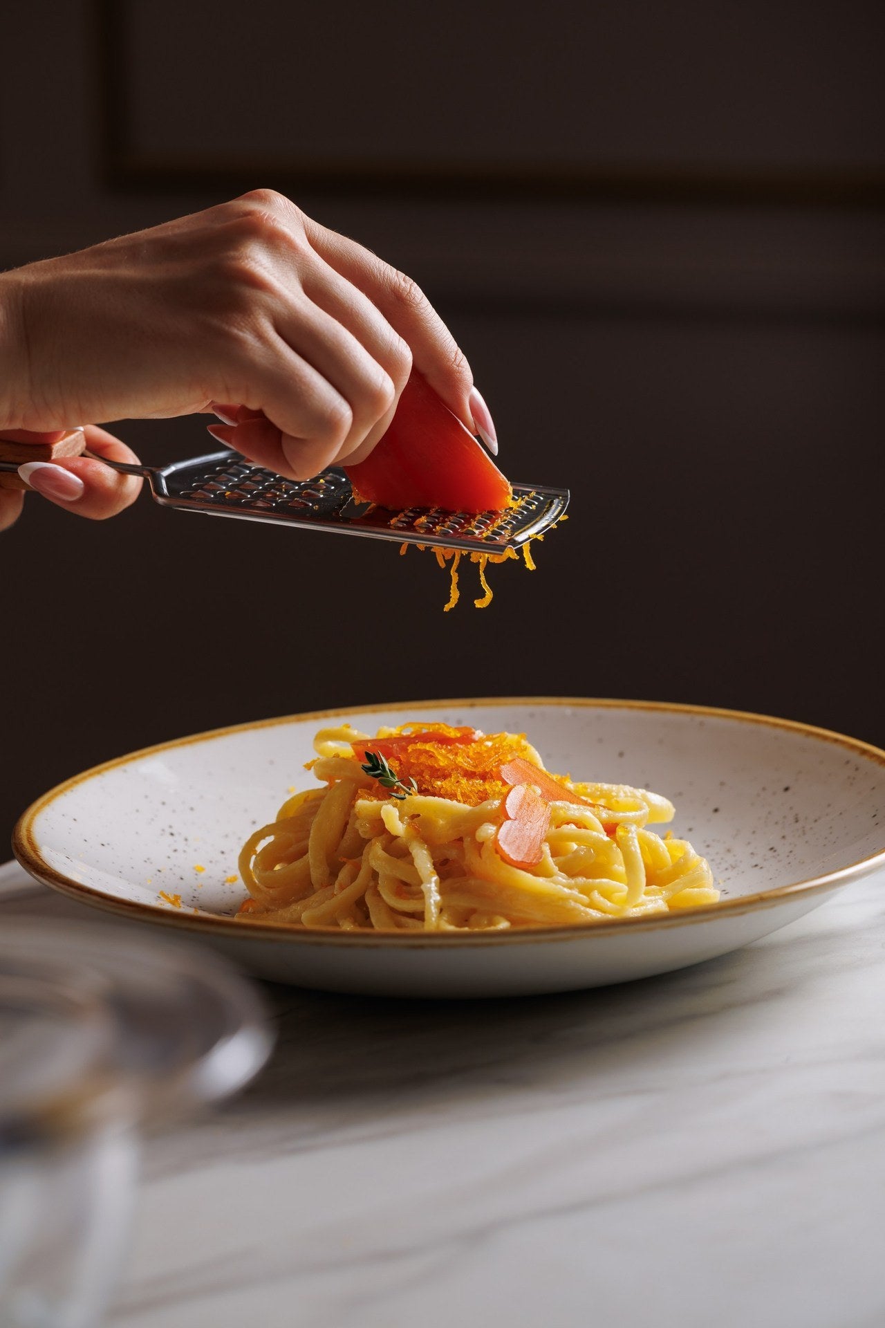 Two hands grating bottarga over a plate of pasta on a white marble table, fine orange strands falling from the grater, against a dark wall.